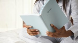 Girl wearing pajamas sitting in bed while holding a blue book that has 'journal' written on the cover