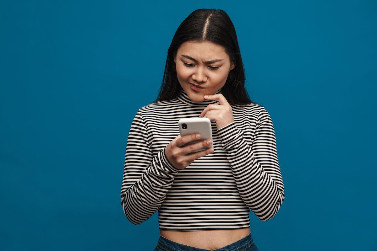 Woman in striped shirt frowning down at phone: Shutterstock. Portrait of a frowning pensive casual young asian woman standing over blue wall background holding mobile phone